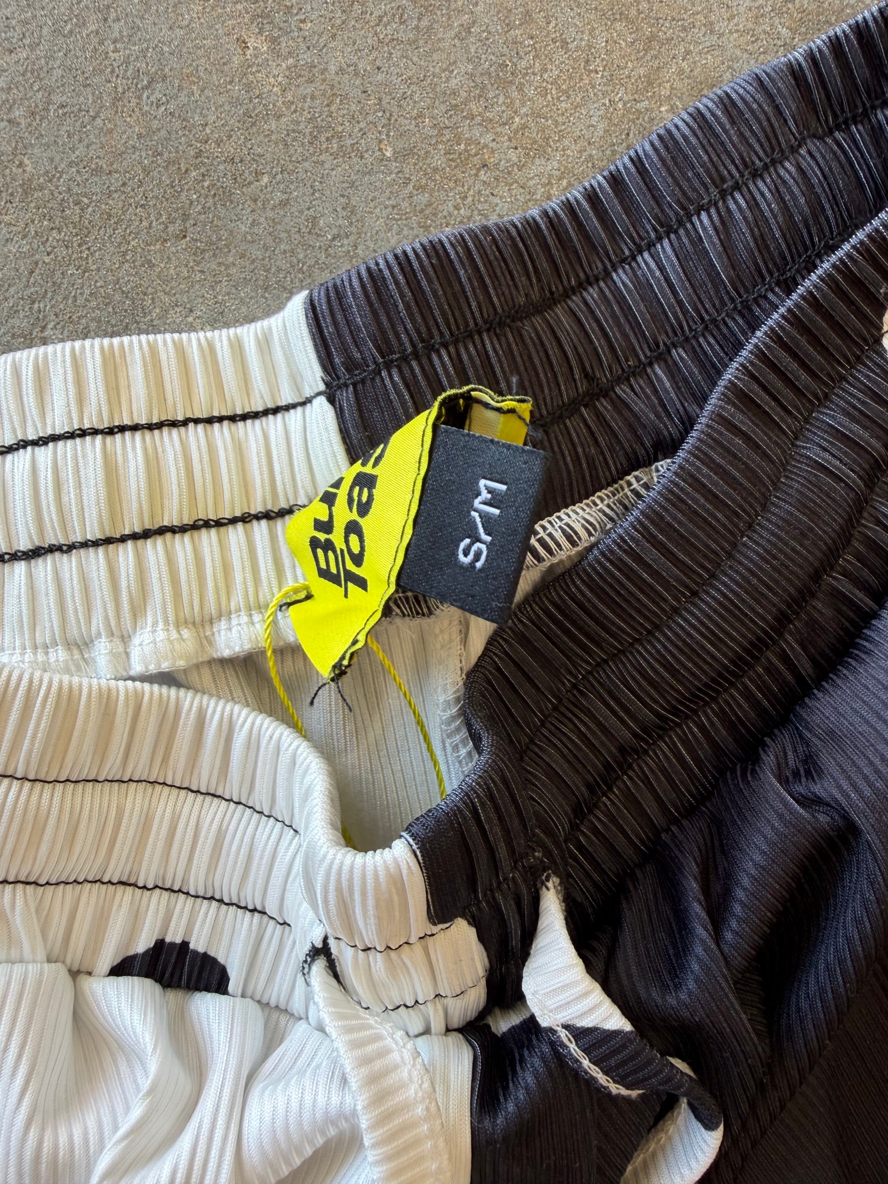 Close-up of pleated athletic shorts in multiple colors, including black, white, and dark gray, with a yellow and black size tag reading S/M
