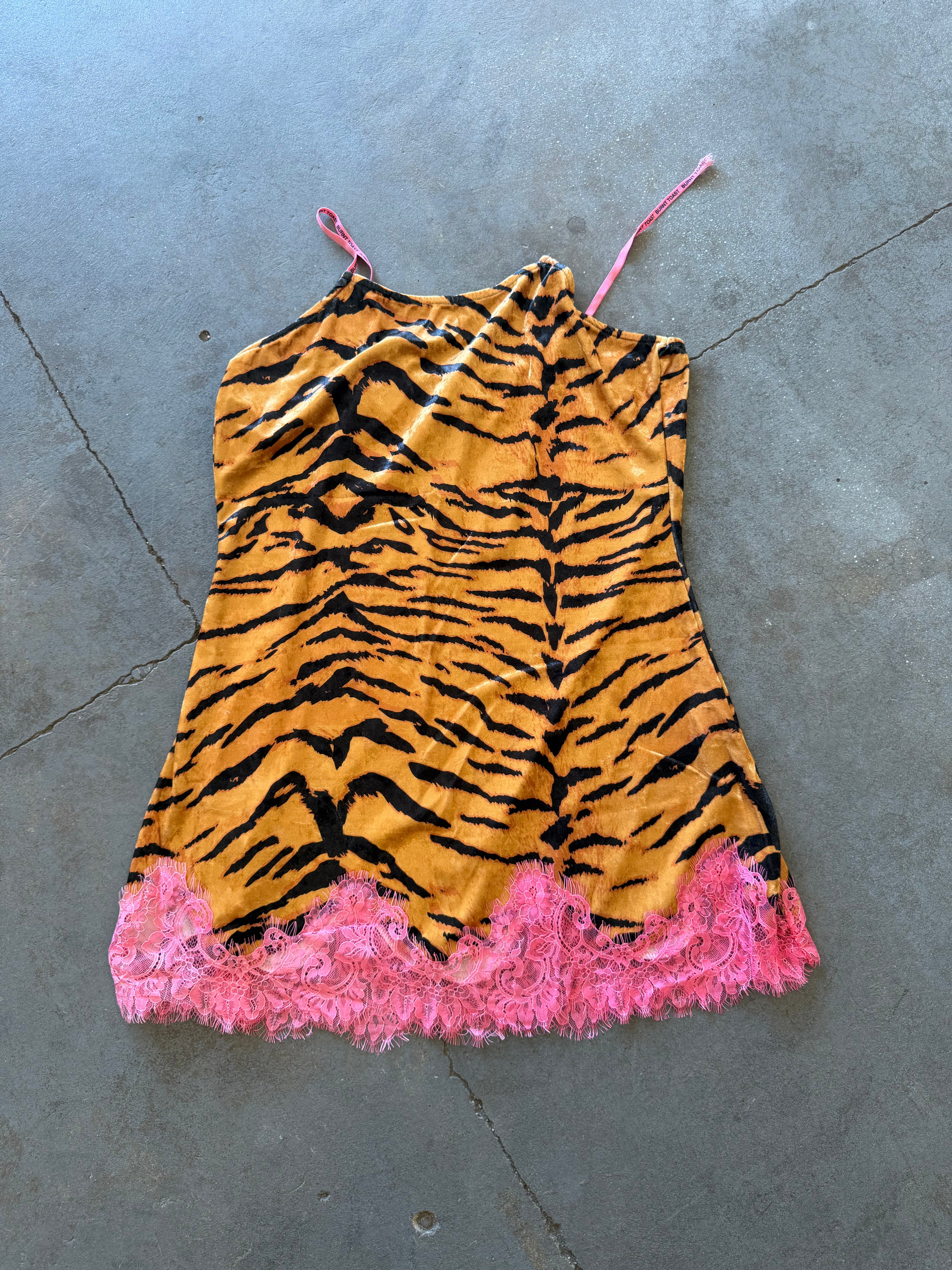 A sleeveless camisole-style top with a tiger-stripe print in orange and black, featuring thin pink straps and a bright pink fringed hem, laid flat on a gray concrete floor