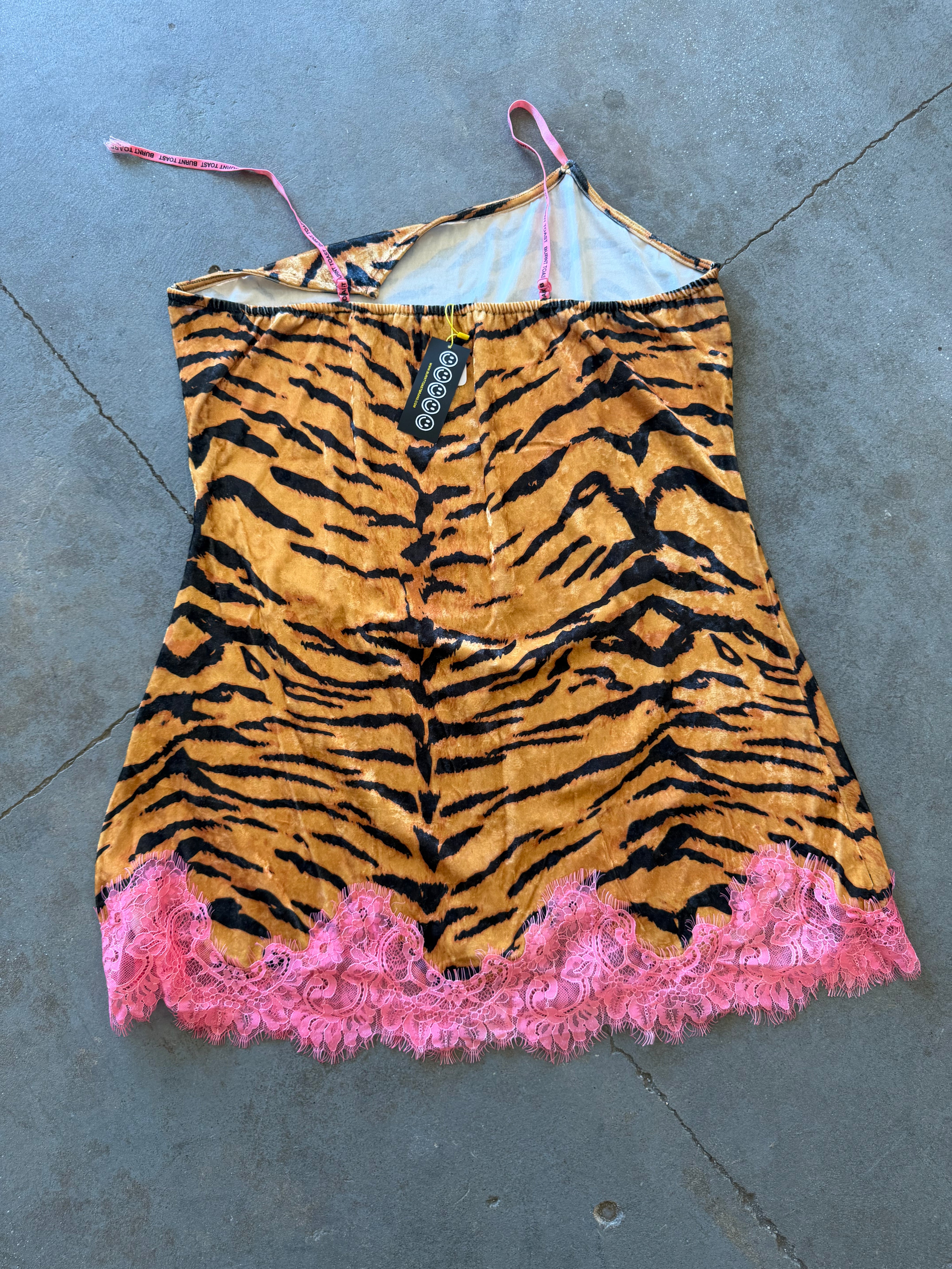 A tiger-print camisole dress with pink lace trim and pink-and-white striped straps, laid flat on a gray concrete floor. A black hangtag with the brand name 'Savage' is visible