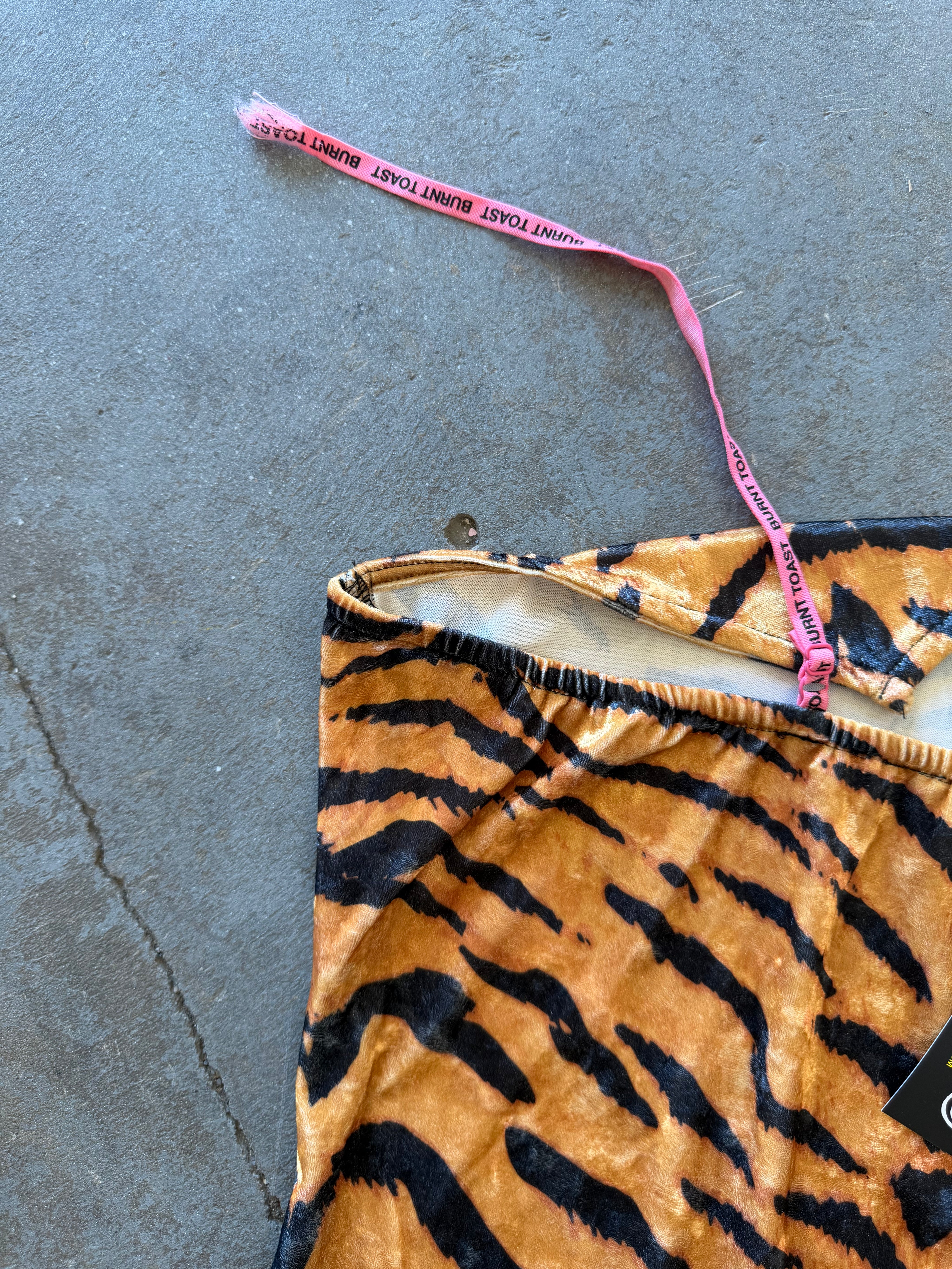 A close-up of a tiger-print fabric item laid on a gray concrete floor, with a pink measuring tape strap draped across it. The fabric has an orange-brown base with bold black stripes and a small portion of a black price tag is visible at the bottom right