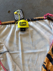 Close-up of a garment laid flat on a gray surface, showing a cream-colored fabric body with a bright yellow Burnt Toast brand label, black elastic trim, and patterned orange and black fabric edges