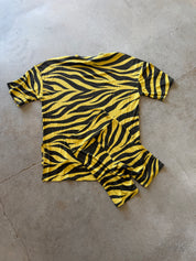 A short-sleeve t-shirt with a bold yellow and black zebra stripe pattern, laid flat on a gray concrete floor