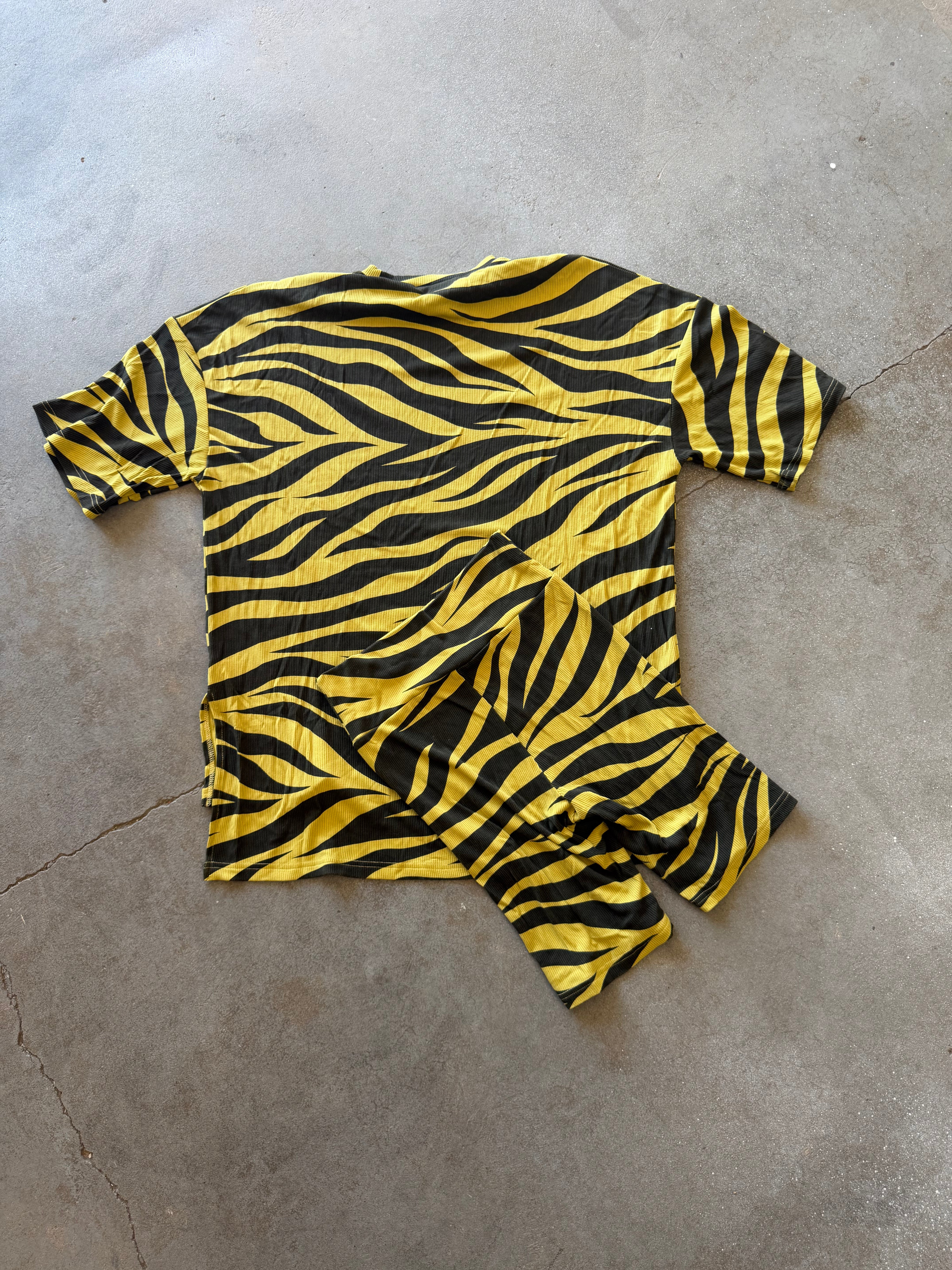 A short-sleeve t-shirt with a bold yellow and black zebra stripe pattern, laid flat on a gray concrete floor