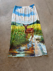 A long A-line skirt with an all-over scenic print of a deer in a meadow under a blue sky, laid flat on a concrete floor