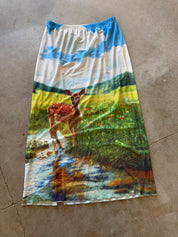 A long A-line skirt with an all-over scenic print of a deer in a meadow under a blue sky, laid flat on a concrete floor, back view