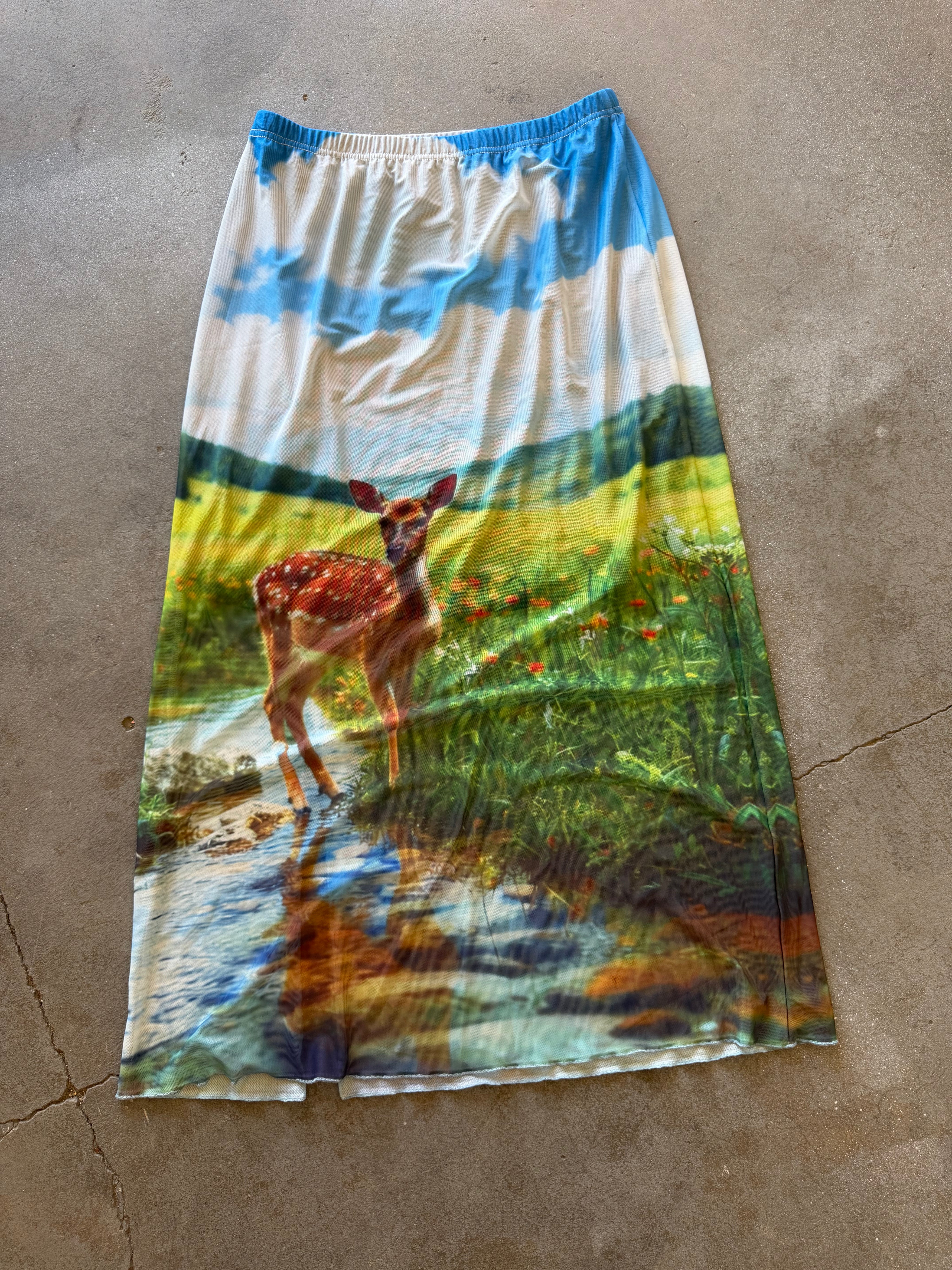A long A-line skirt with an all-over scenic print of a deer in a meadow under a blue sky, laid flat on a concrete floor, back view