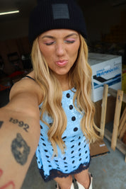 A woman taking a selfie in a garage-like setting, wearing a black beanie with a 'sugar wash' patch and a light blue sleeveless dress with black polka dots