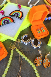 A styled flat-lay of colorful handmade jewelry, including rainbow beaded earrings, patterned hoop earrings, a green bead necklace, and paintbrush-shaped earrings, arranged around orange jewelry boxes on a dark textured surface