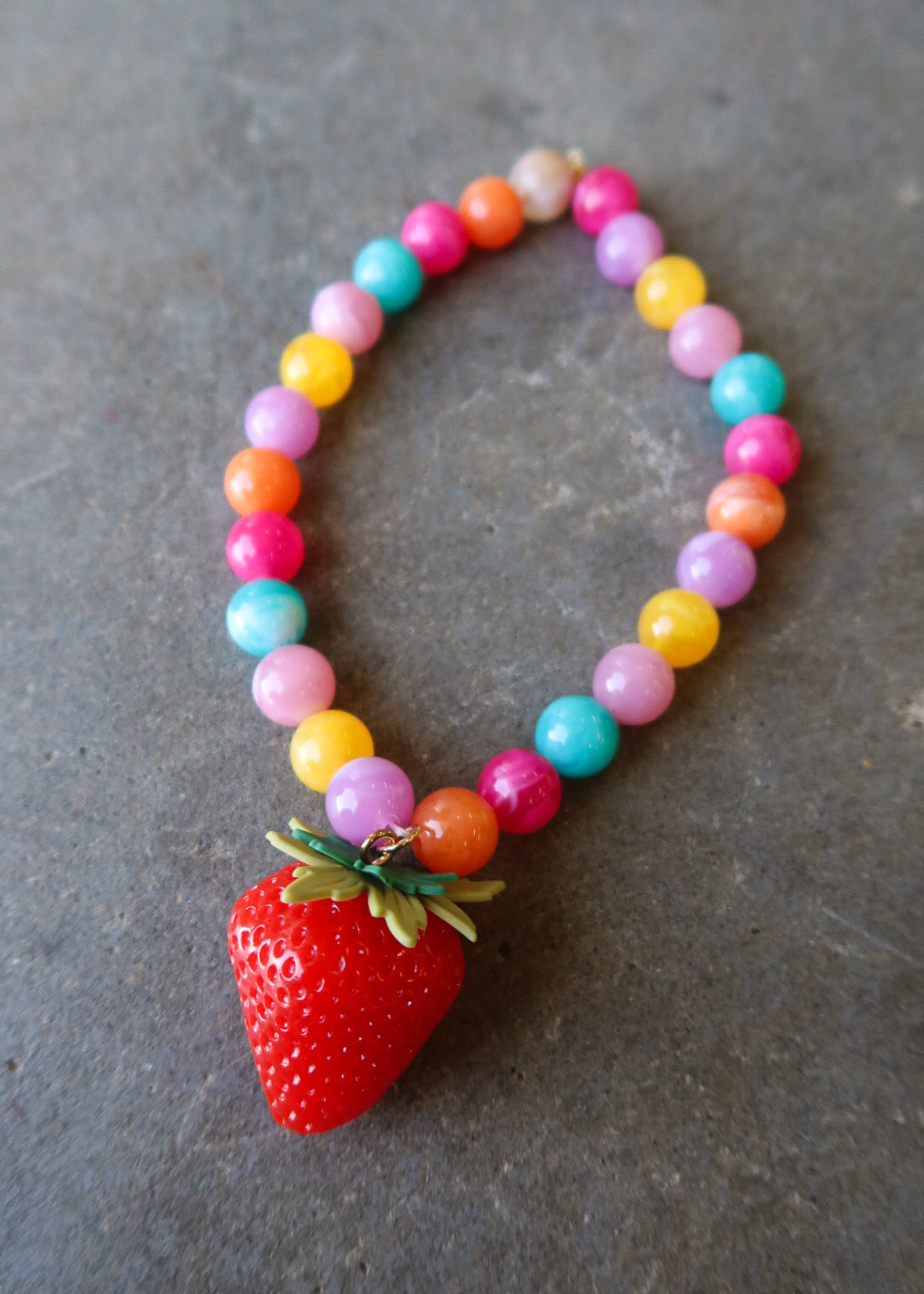 A colorful beaded necklace with a red strawberry charm, displayed on a textured gray surface