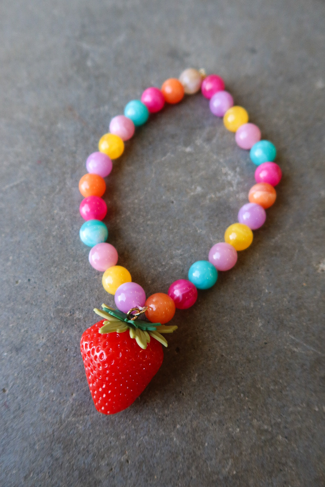 A colorful beaded necklace with a red strawberry charm, displayed on a textured gray surface