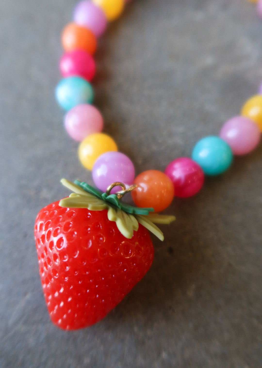 A close-up of a colorful beaded necklace with a large red strawberry pendant, resting on a dark gray textured surface