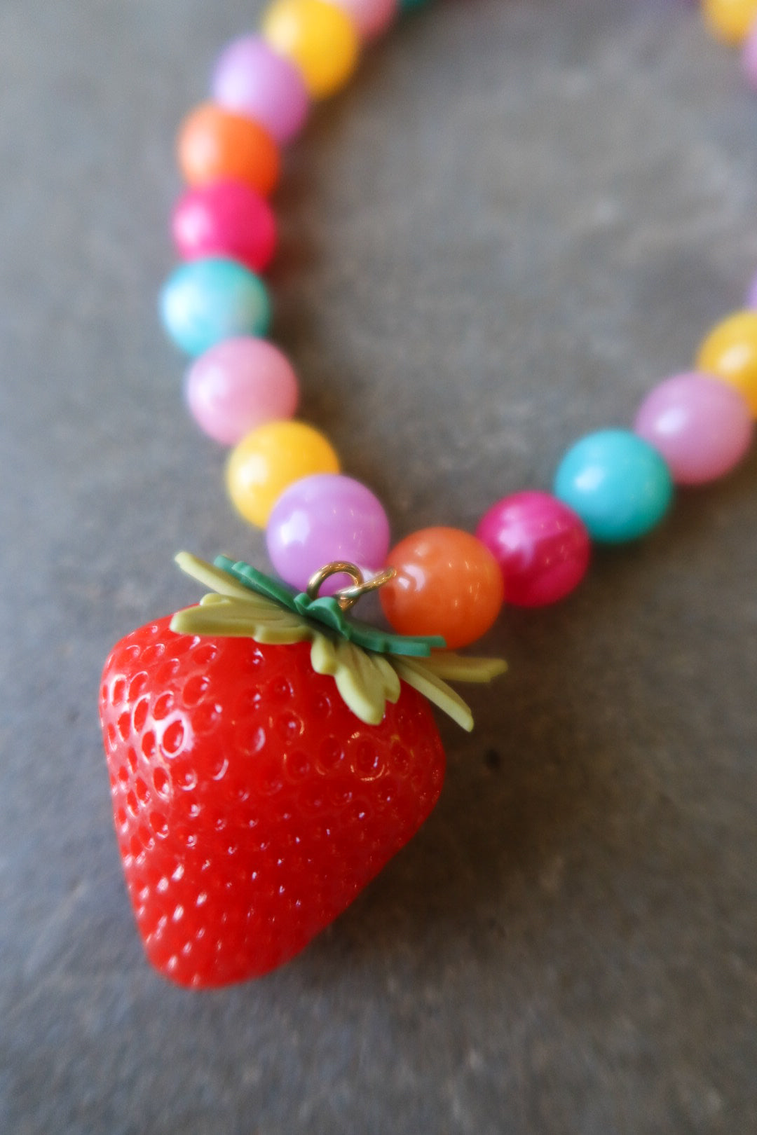 A close-up of a colorful beaded necklace with a large red strawberry pendant, resting on a dark gray textured surface