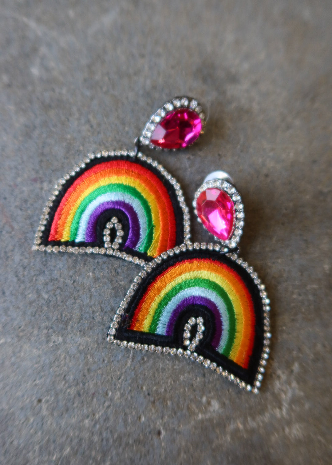 A pair of rainbow embroidered patch-style earrings with silver-tone rhinestone borders and bright pink teardrop studs, displayed on a textured gray surface
