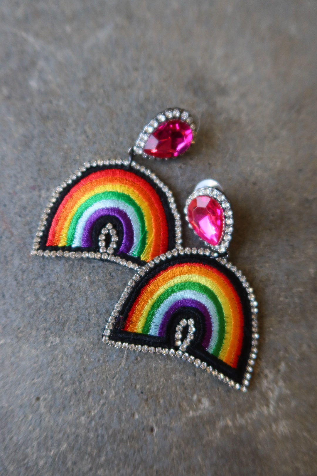 A pair of rainbow embroidered patch-style earrings with silver-tone rhinestone borders and bright pink teardrop studs, displayed on a textured gray surface