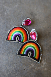 A pair of rainbow embroidered patch-style earrings with silver-tone rhinestone borders and bright pink teardrop studs, displayed on a textured gray surface