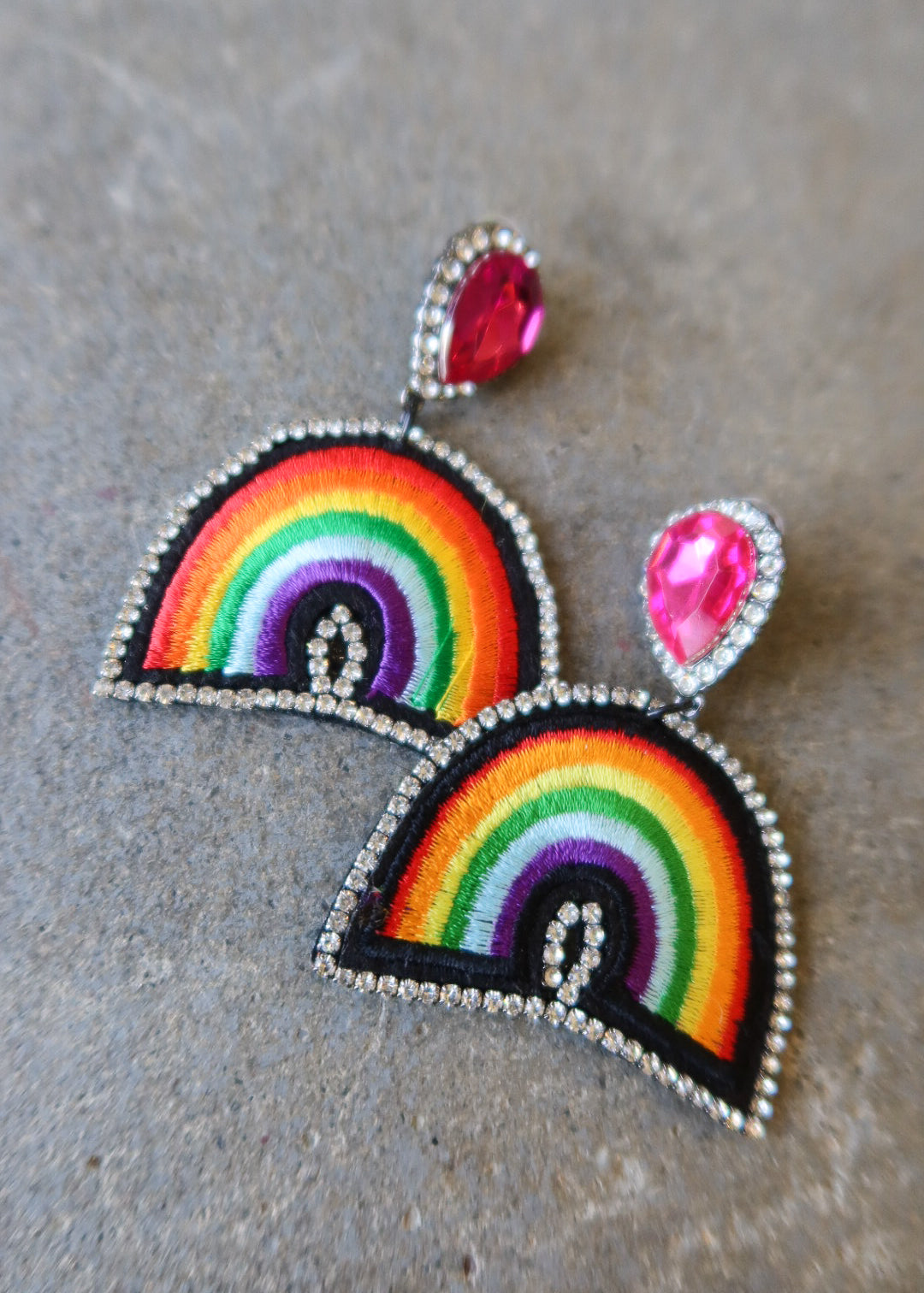 A pair of handmade rainbow embroidered patchwork earrings with silver rhinestone borders and pink teardrop studs, displayed on a textured gray surface