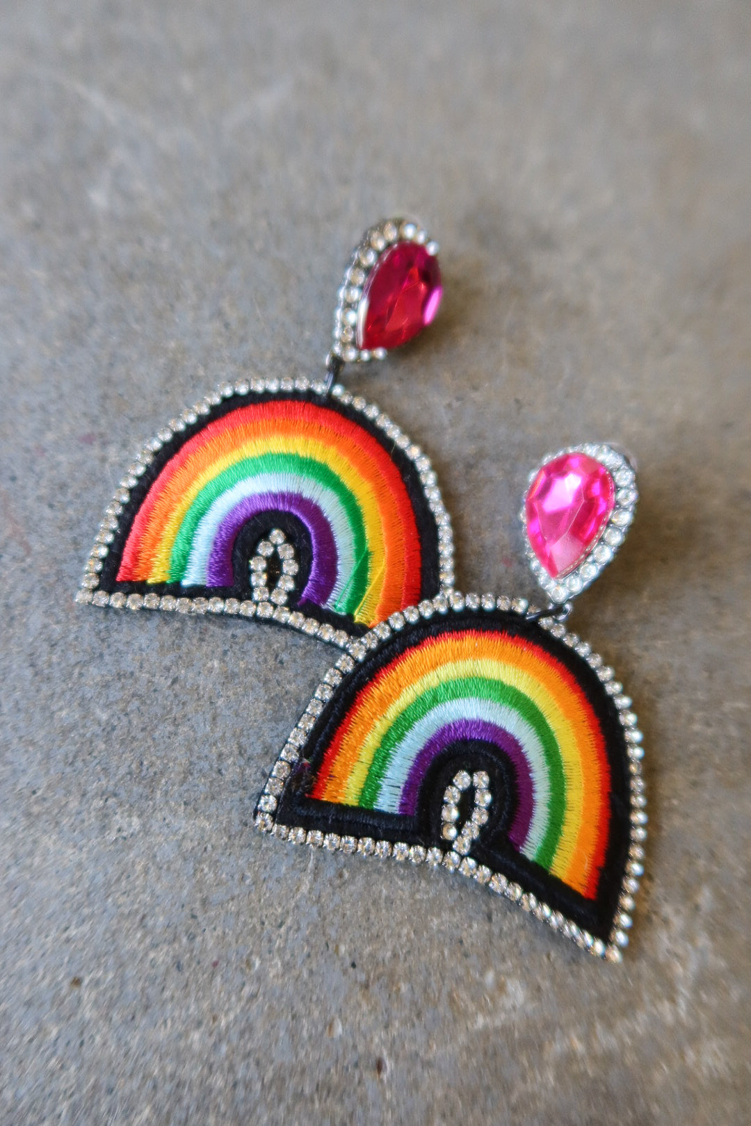 A pair of handmade rainbow embroidered patchwork earrings with silver rhinestone borders and pink teardrop studs, displayed on a textured gray surface