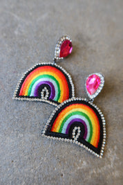 A pair of handmade rainbow embroidered patchwork earrings with silver rhinestone borders and pink teardrop studs, displayed on a textured gray surface