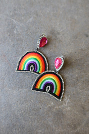 A pair of rainbow arch drop earrings with silver-tone metal borders, pink teardrop studs, and small clear rhinestone accents, displayed on a textured gray surface