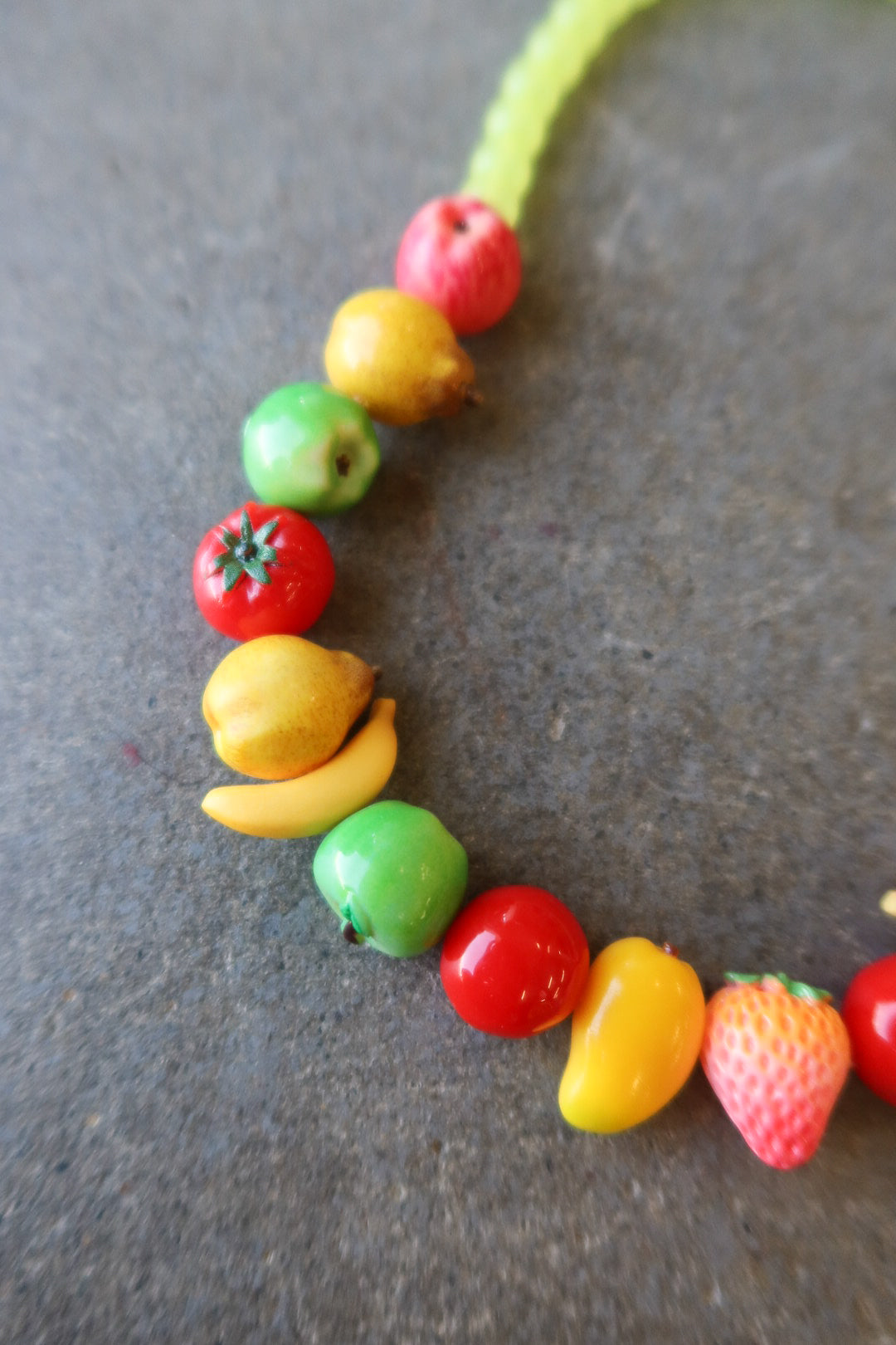A novelty necklace made of small, colorful fruit-shaped beads, including red strawberries, yellow pears, green apples, and red tomatoes, laid out on a textured gray surface