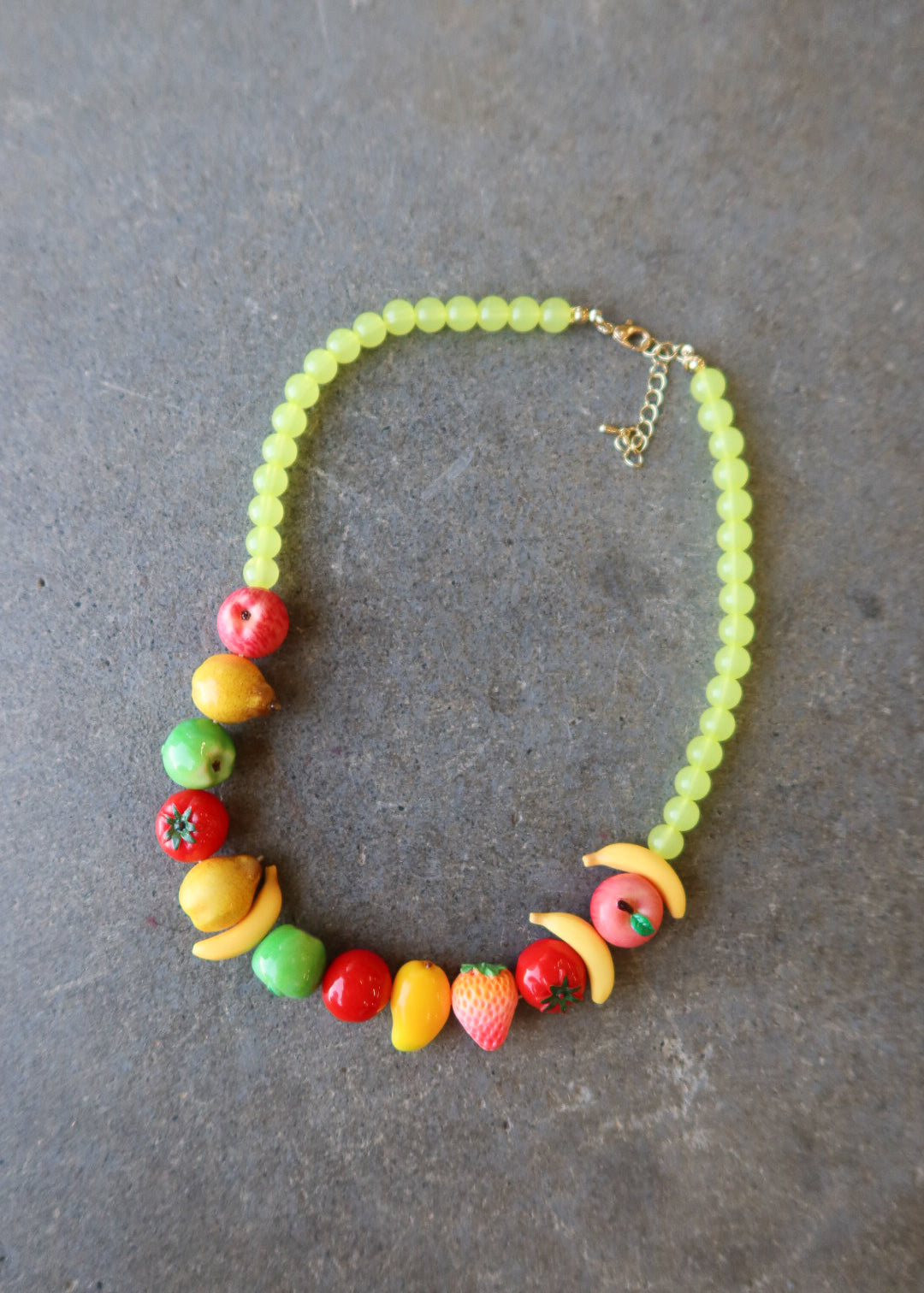 A beaded necklace featuring light green round beads and colorful fruit-shaped accents in red, yellow, orange, and green, displayed on a dark gray textured surface