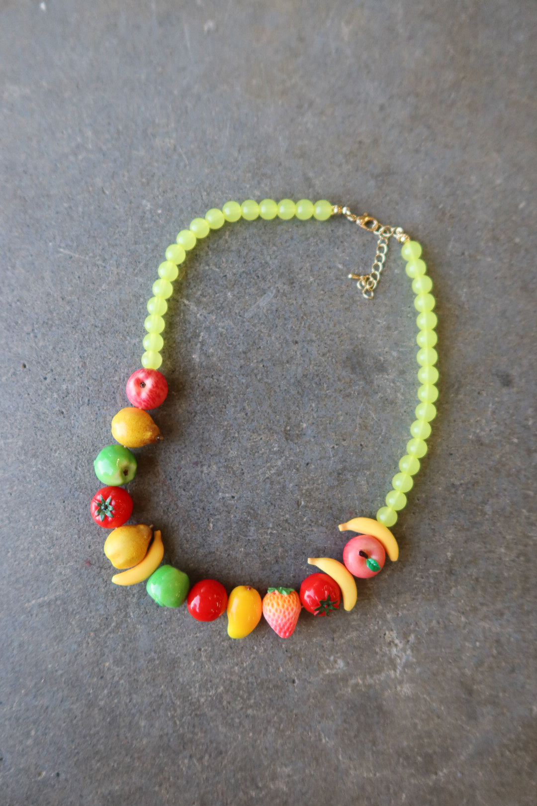 A beaded necklace featuring light green round beads and colorful fruit-shaped accents in red, yellow, orange, and green, displayed on a dark gray textured surface