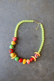 A beaded necklace featuring light green round beads and colorful fruit-shaped accents in red, yellow, orange, and green, displayed on a dark gray textured surface