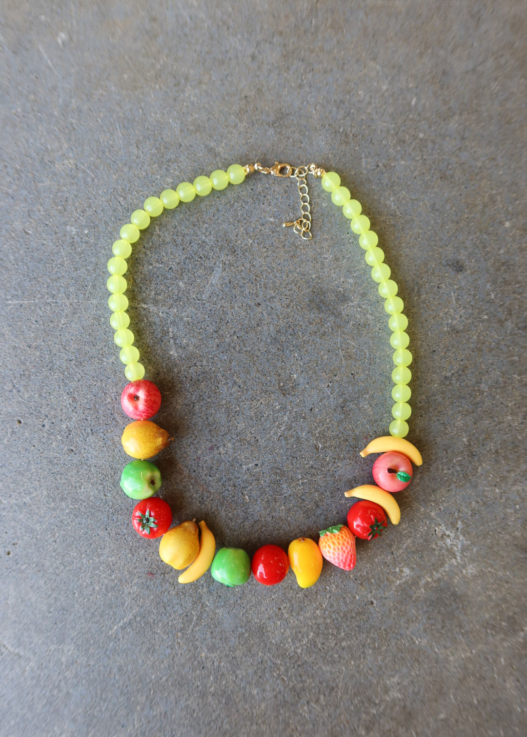A beaded necklace with light green round beads and colorful fruit-shaped charms, displayed on a textured gray background