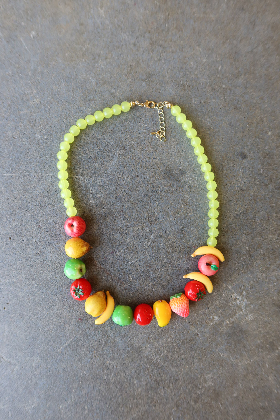 A beaded necklace with light green round beads and colorful fruit-shaped charms, displayed on a textured gray background