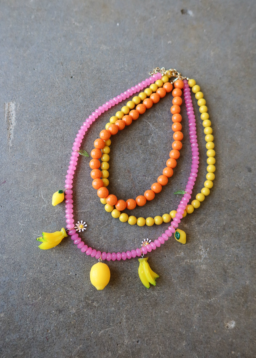 Three beaded necklaces in pink, orange, and yellow, styled with small yellow banana and lemon charms, displayed on a gray concrete surface