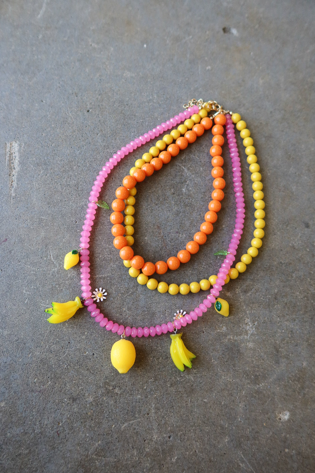 Three beaded necklaces in pink, orange, and yellow, styled with small yellow banana and lemon charms, displayed on a gray concrete surface