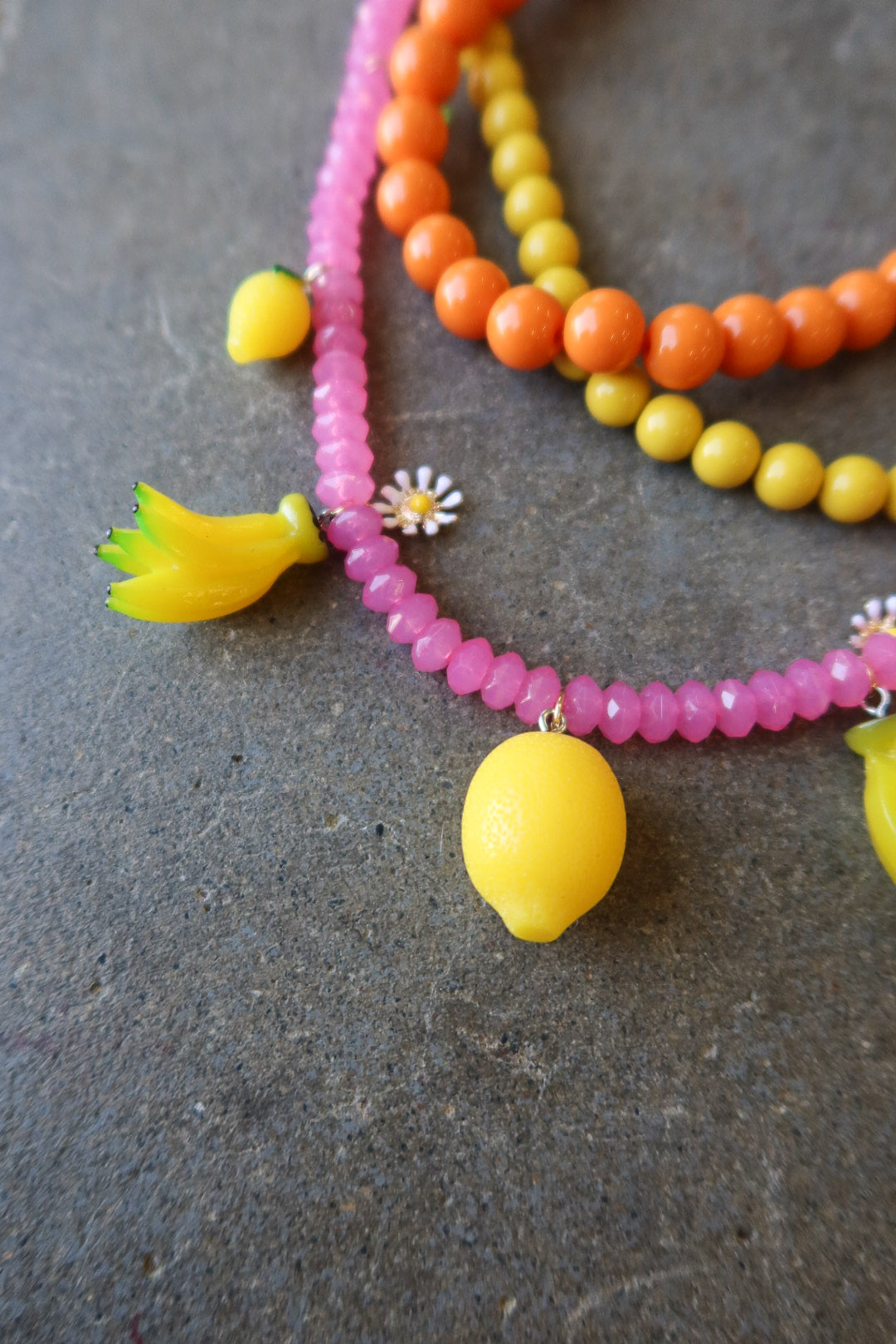 A close-up of a beaded necklace featuring bright pink faceted beads, orange and yellow round beads, and yellow lemon-shaped charms, laid on a dark textured surface