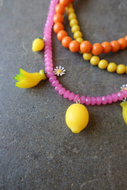 A close-up of a beaded necklace featuring bright pink faceted beads, orange and yellow round beads, and yellow lemon-shaped charms, laid on a dark textured surface