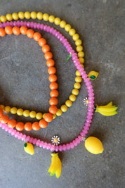 Three beaded necklaces in pink, orange, and yellow, decorated with small yellow banana and lemon charms, displayed on a textured gray surface