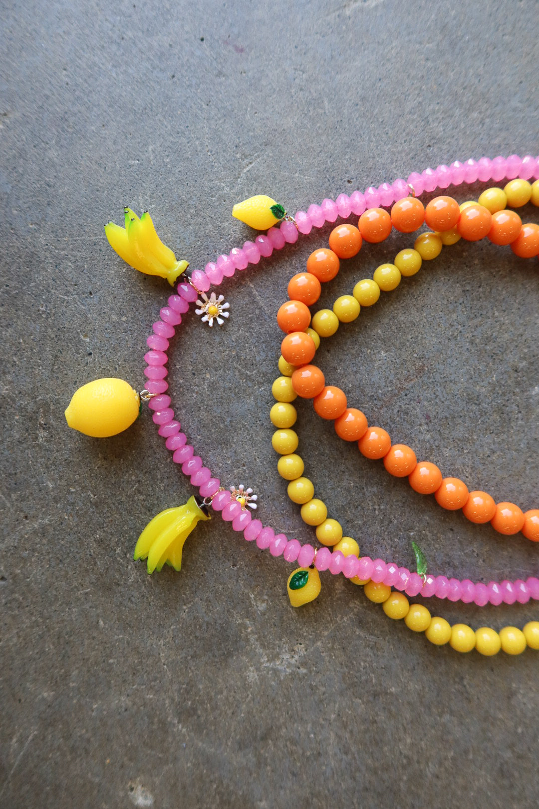 Three beaded strands of necklaces in pink, orange, and yellow, decorated with small fruit charms including lemons and bananas, arranged on a textured gray surface