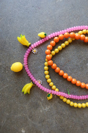 Three beaded strands of necklaces in pink, orange, and yellow, decorated with small fruit charms including lemons and bananas, arranged on a textured gray surface