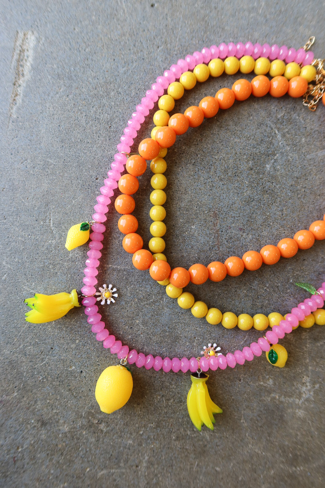 Three beaded necklaces in pink, orange, and yellow, decorated with small fruit charms and flower details, arranged on a gray stone surface