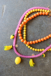 Three beaded necklaces in pink, orange, and yellow, decorated with small fruit charms and flower details, arranged on a gray stone surface