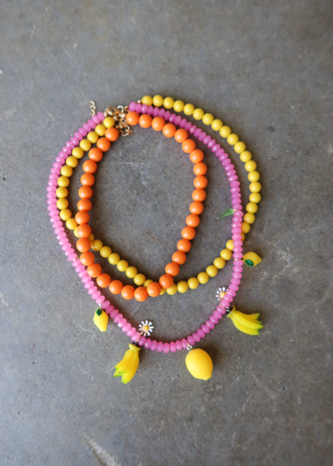 Three beaded necklaces in pink, orange, and yellow, styled with small yellow flower and banana charms, arranged on a gray concrete surface