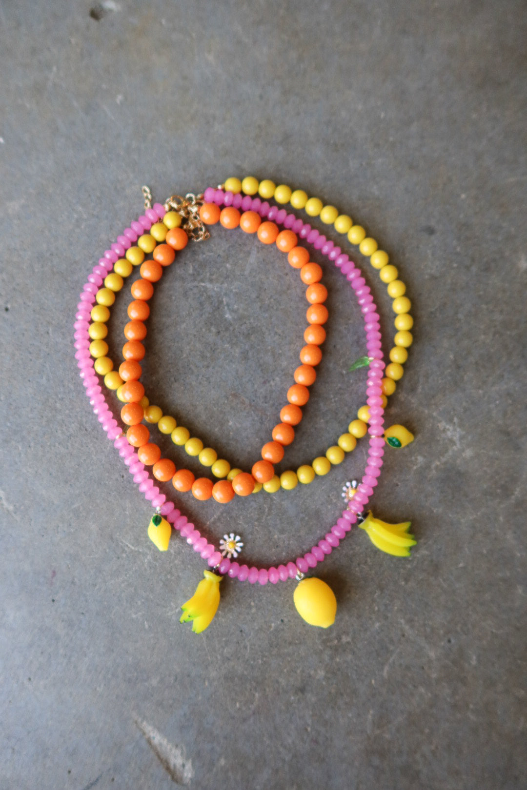 Three beaded necklaces in pink, orange, and yellow, styled with small yellow flower and banana charms, arranged on a gray concrete surface