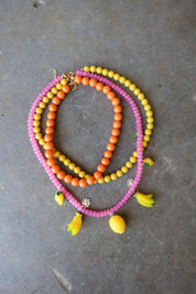 Three beaded necklaces in pink, orange, and yellow, styled with small yellow flower and banana charms, arranged on a gray concrete surface