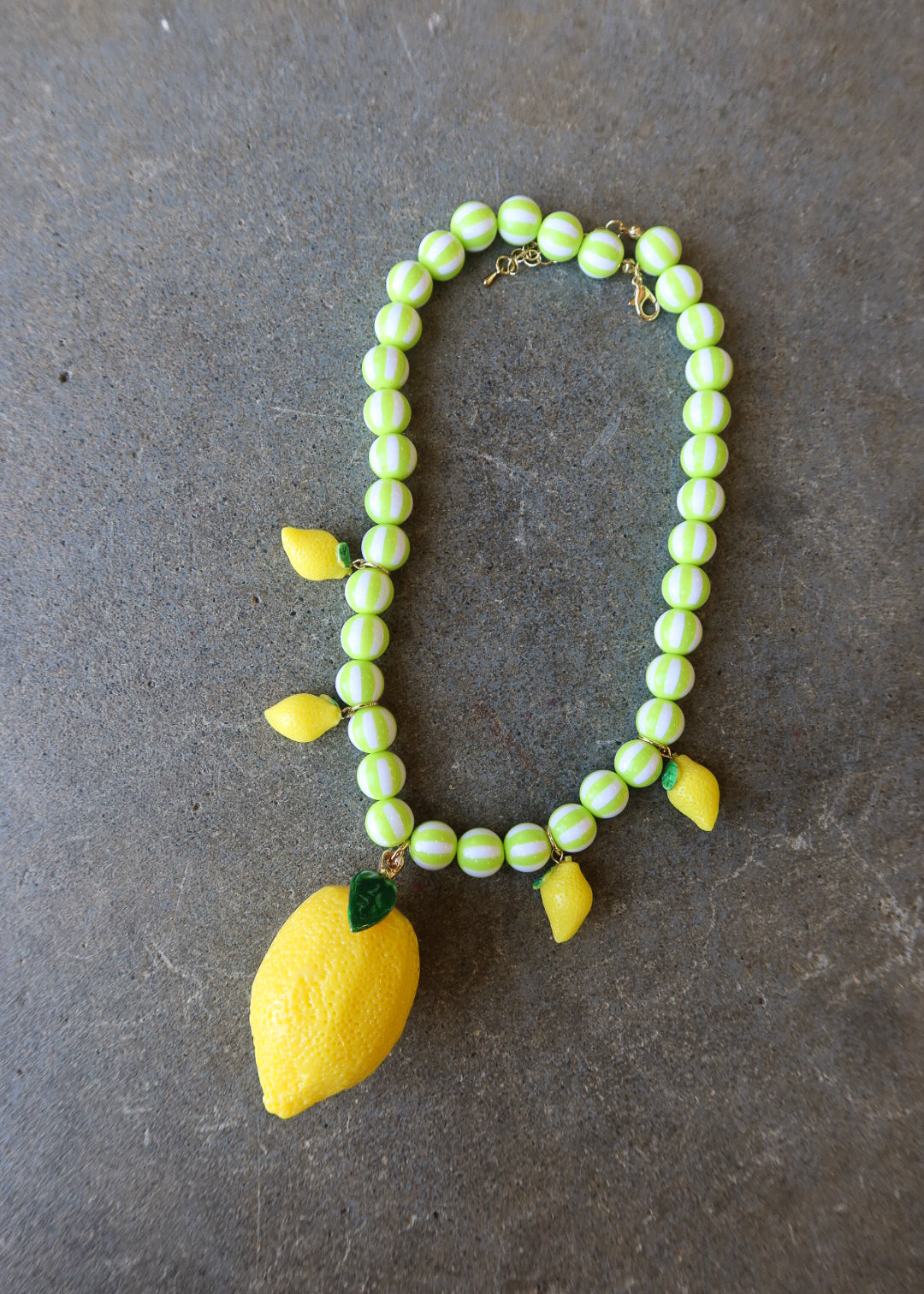 A beaded necklace featuring light green and white striped beads, yellow lemon-shaped charms, and a large yellow lemon pendant with a green leaf, displayed on a dark gray textured surface