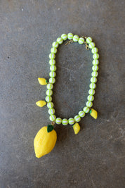 A beaded necklace featuring light green and white striped beads, yellow lemon-shaped charms, and a large yellow lemon pendant with a green leaf, displayed on a dark gray textured surface