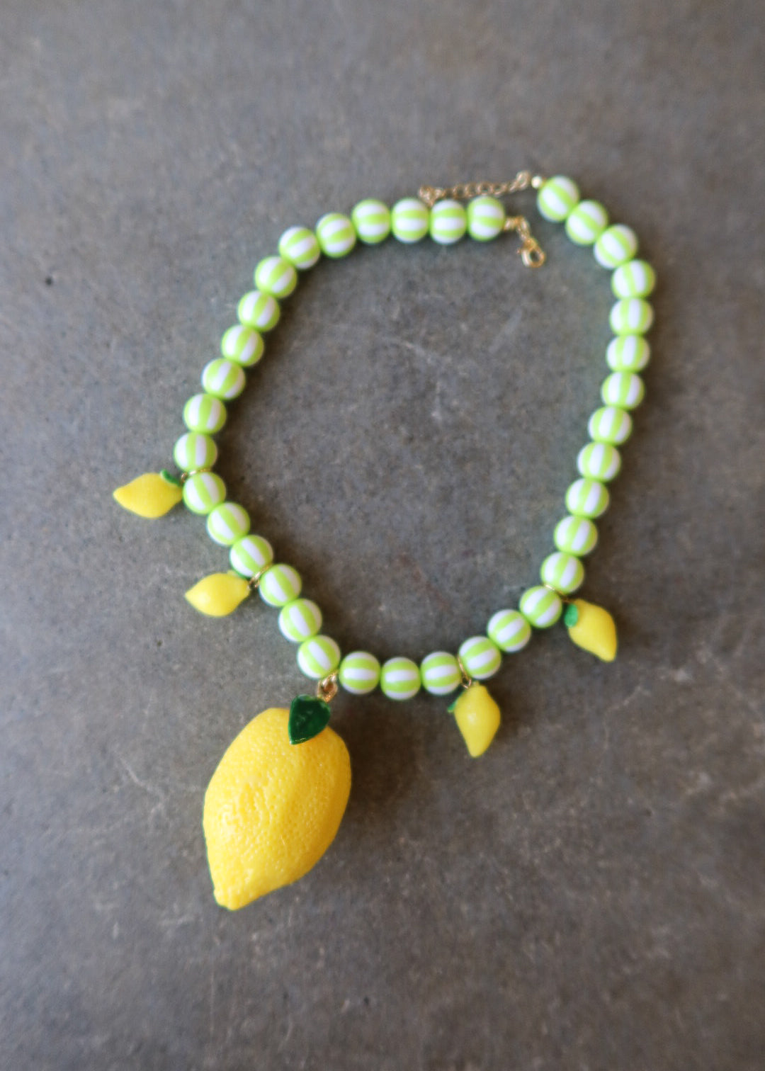 A beaded necklace featuring light green and white striped beads, yellow lemon charms, and a large yellow lemon pendant with green leaves, displayed on a dark gray textured surface
