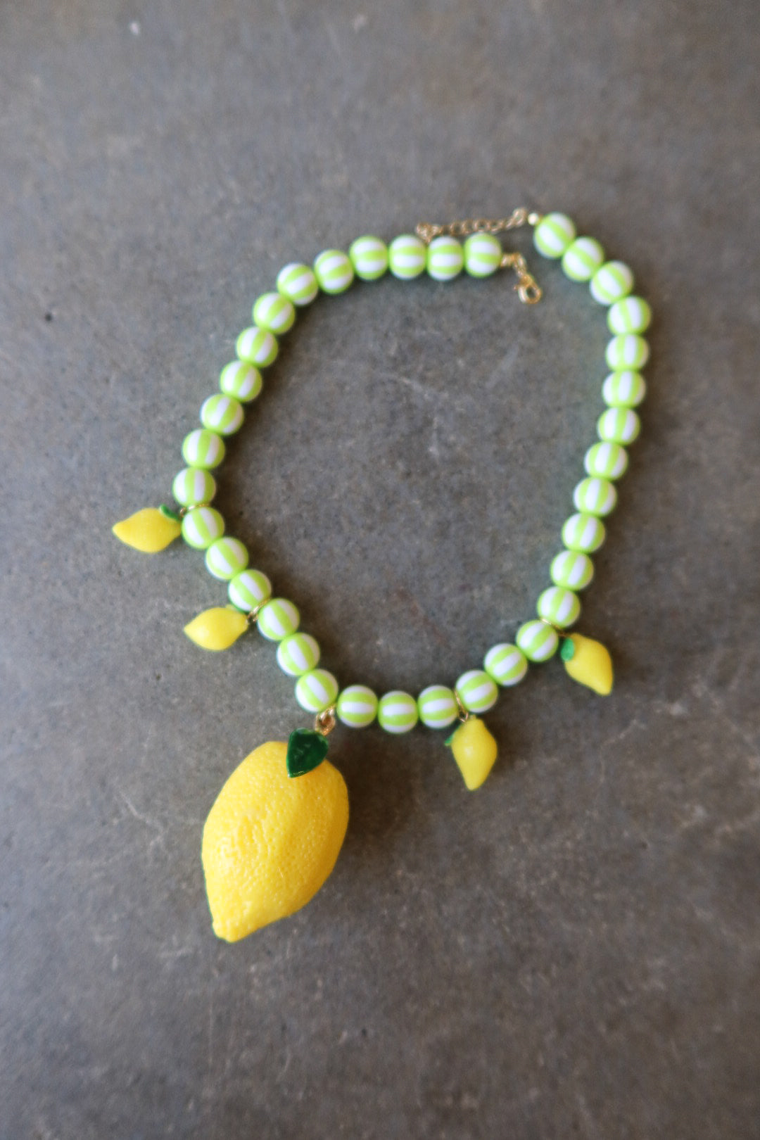 A beaded necklace featuring light green and white striped beads, yellow lemon charms, and a large yellow lemon pendant with green leaves, displayed on a dark gray textured surface