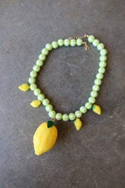 A beaded necklace featuring light green and white striped beads, yellow lemon charms, and a large yellow lemon pendant with green leaves, displayed on a dark gray textured surface
