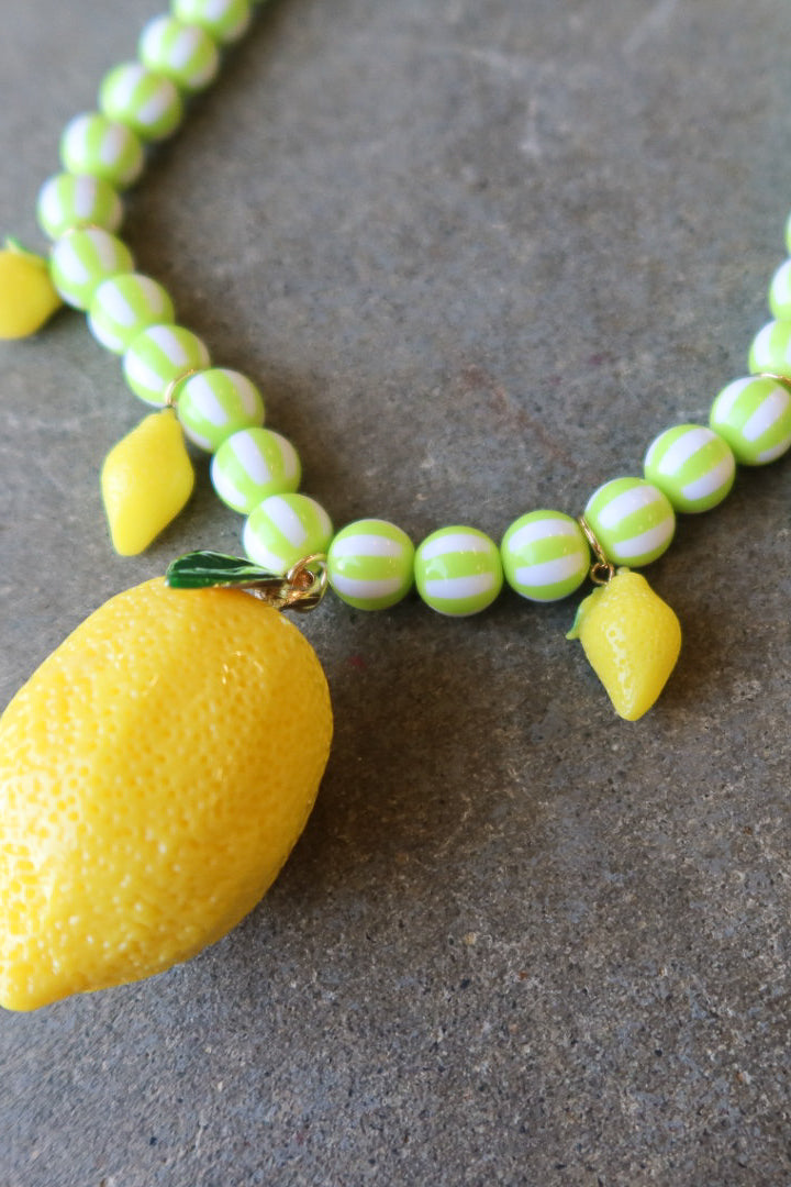 A beaded necklace featuring light green and white striped round beads, small yellow bead accents, and a large yellow lemon-shaped pendant, displayed on a textured gray surface
