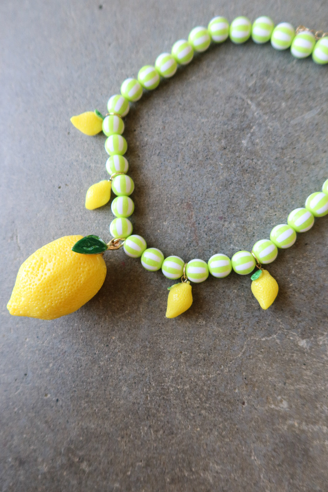 A beaded necklace featuring light green and white striped round beads, yellow lemon charms, and a large textured yellow lemon pendant with a green leaf, displayed on a gray stone surface