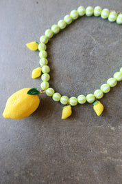 A beaded necklace featuring light green and white striped round beads, yellow lemon charms, and a large textured yellow lemon pendant with a green leaf, displayed on a gray stone surface