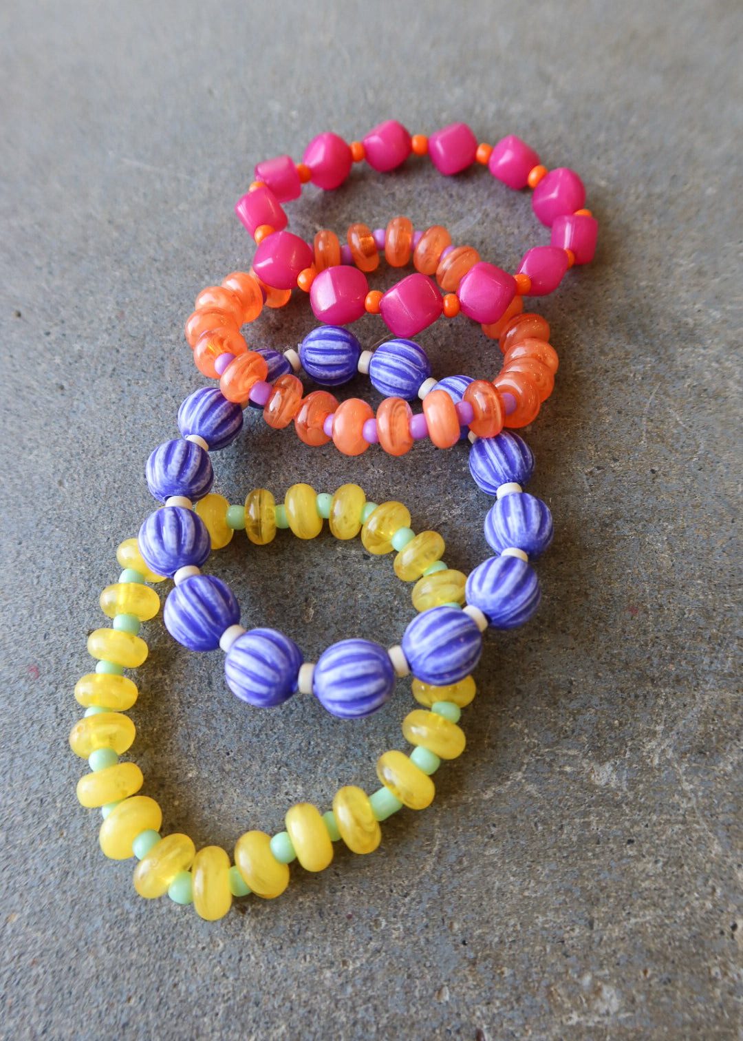 Four beaded stretch bracelets in pink, orange, purple, and yellow, arranged on a textured gray surface
